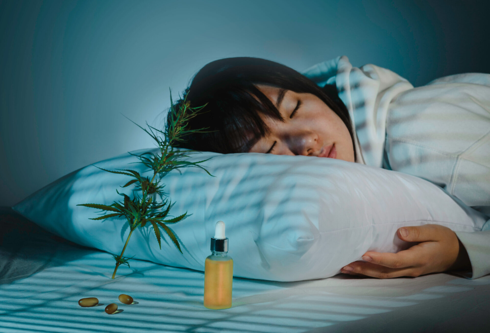 夕方の寝室で眠っている女性のそばに、CBDオイルやカプセル、カンナビスの枝が置かれ、睡眠やメラトニン分泌を表現した様子